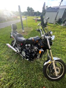 A black 1983 Honda Magna motorcycle parked on grass in the sunlight.