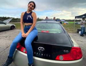 A woman smiling, seated on the trunk of a black Infiniti G35 at Orlando Speed World in 2022, with other cars and spectators in the background.